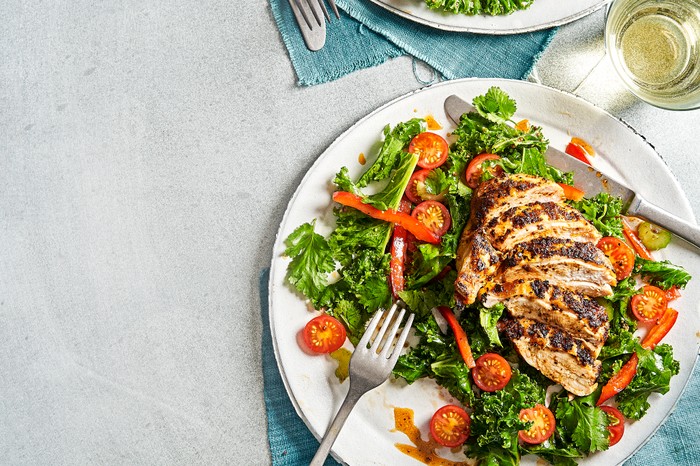 Cajun chicken supreme A grey background with two white plates topped with green salad leaves, halved tomatoes and slices of cooked chicken
