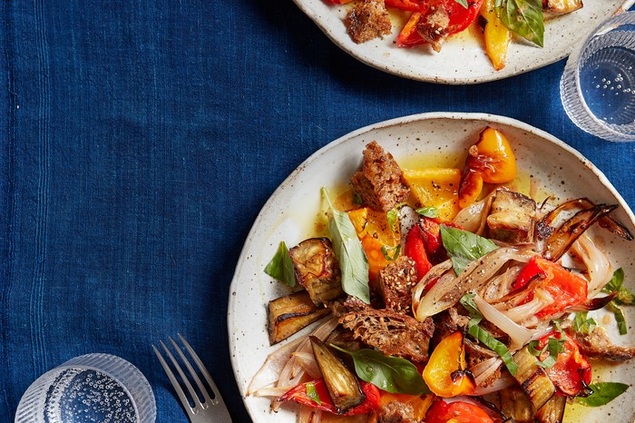 Healthy panzanella A blue tablecloth topped with two grey plates. Each plate is filled with red and yellow roasted vegetable and green basil leaves