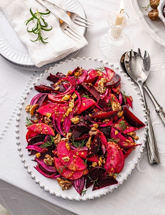 A sharing beetroot salad on a white plate with serving cutlery on the side