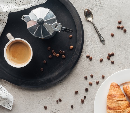 top view of cup of coffee and moka pot on concrete surface with spilled coffee beans and plate of croissants
