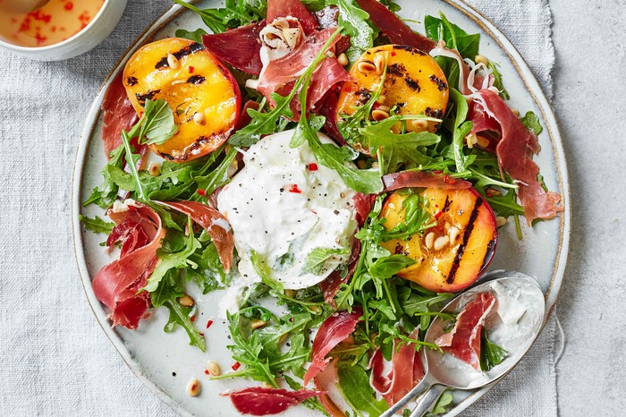 Grilled Peach Salad with Burrata Recipe A grey plate topped with green leaves, orange peaches and a ball of white cheese