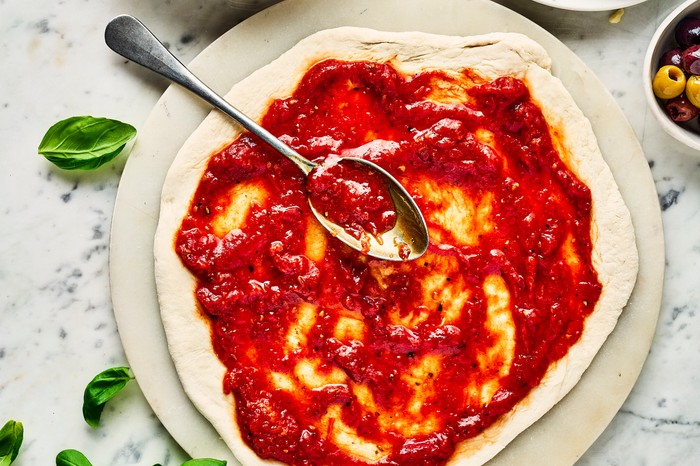 Pizza sauce An uncooked pizza base with tomato sauce spread over it, next to a plate of grated cheese, bowl of more sauce and some fresh basil leaves