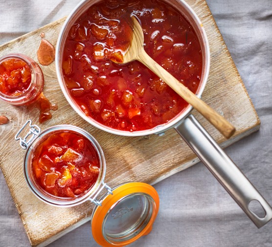 A silver saucepan filled with red jam and a wooden spoon next to a jar of jam on a wooden board