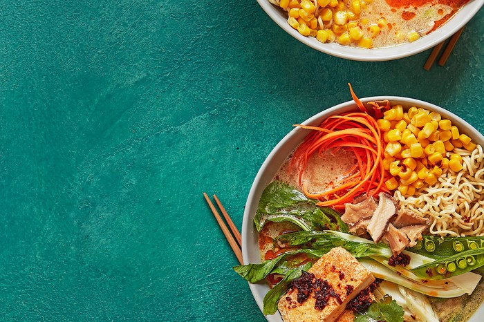 A teal blue background with two bowls of veg-packed ramen