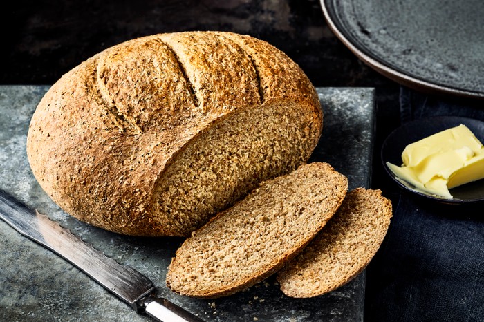 A loaf of wholemeal bread with a couple of slices cut out, on a black board with a knife and knob of butter on the side